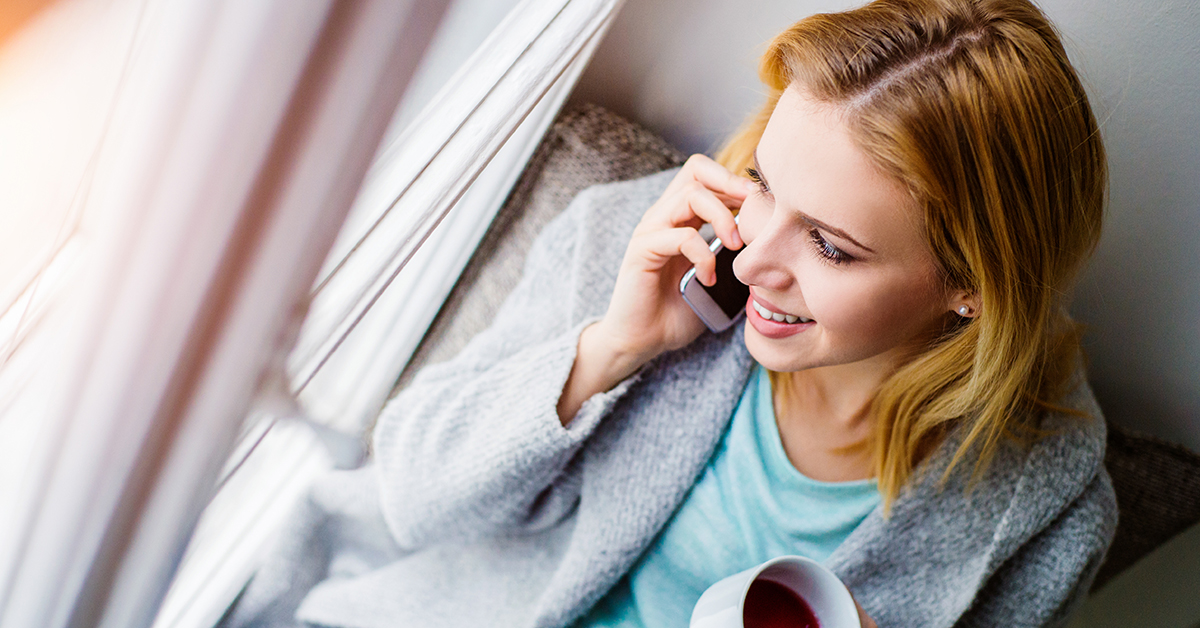 Woman sitting on window sill with smart phone and cup of tea making a phone call.