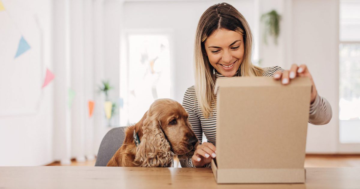 One woman, female opening cardboard boxes package that she just received. Her dog is sitting next to her.