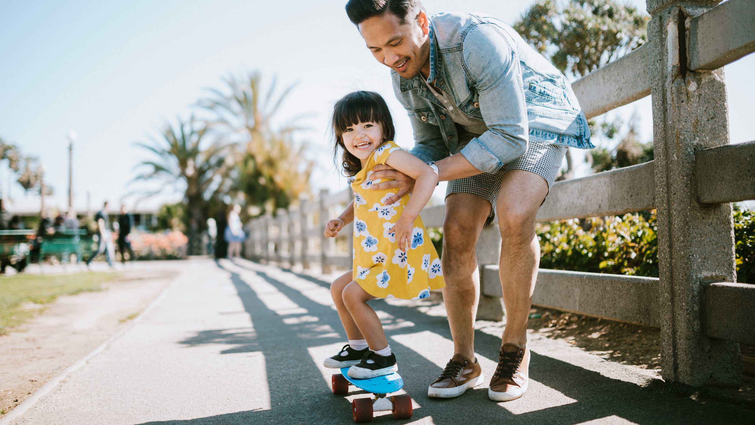 A dad helps his little girl go skateboarding, holding her waist for support. Shot in Los Angeles, California by the Santa Monica Pier.