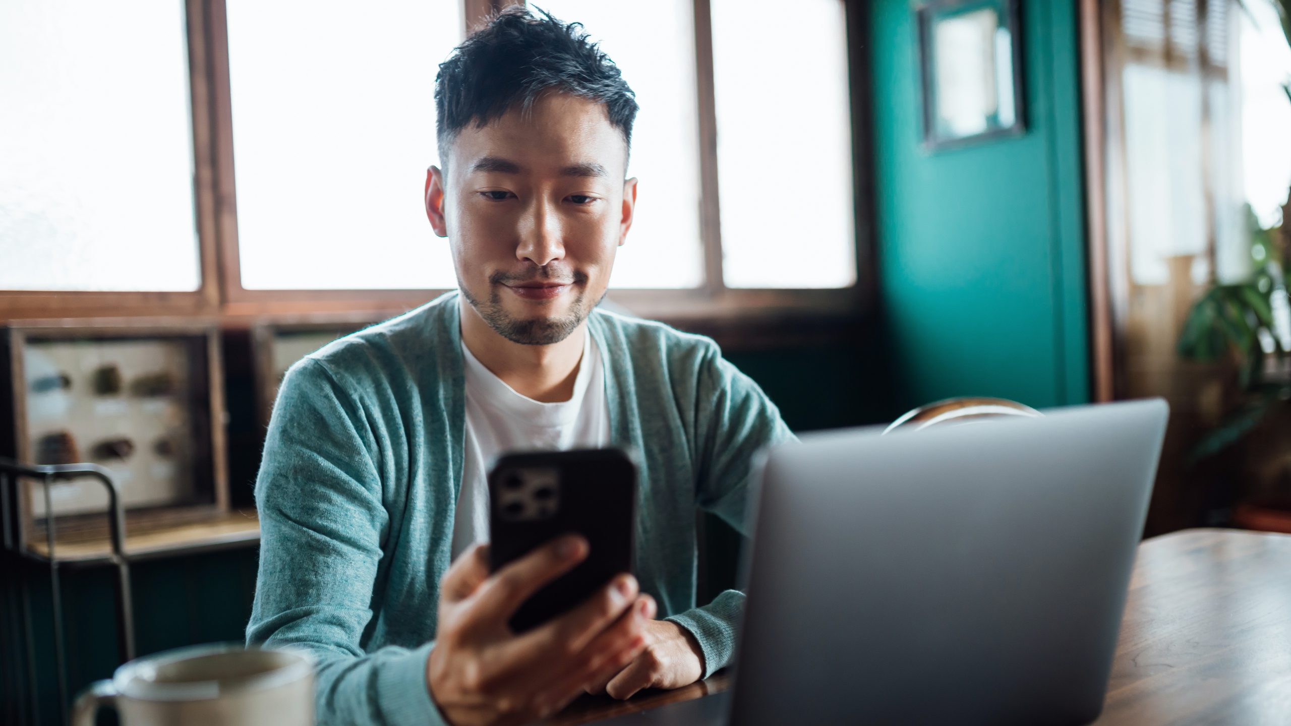 Confident young Asian man looking at smartphone while working on laptop computer in home office. Remote working, freelancer, small business concept
